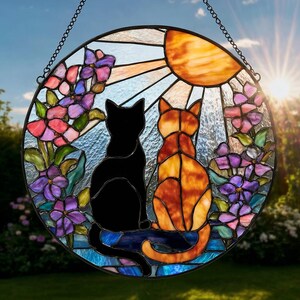 以下が含まれることがあります： 黒猫とオレンジ色のトラ猫が並んで座っている様子を描いた円形のステンドグラスのサンキャッチャー。背景には光を放つ明るいオレンジ色の太陽が描かれ、その周囲には紫やピンクの花の房と緑の葉が配置されています。