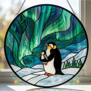 May include: A circular stained glass suncatcher featuring an adult penguin and a chick standing on a snowy landscape. The background depicts a vibrant green and blue aurora borealis in the night sky with small green pine trees on the horizon.