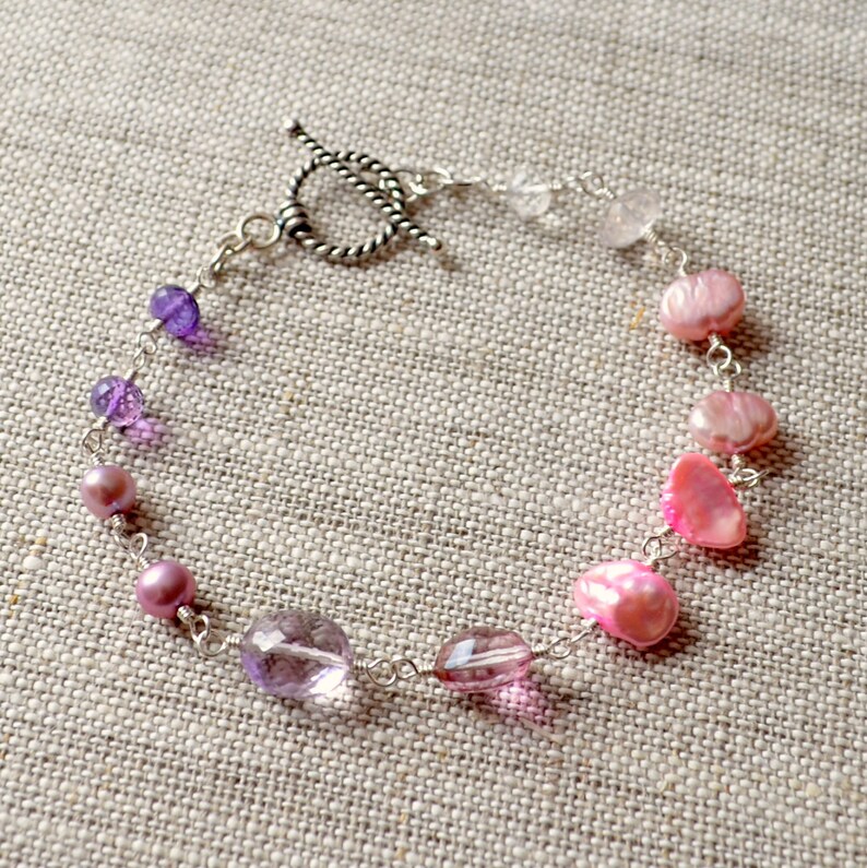 May include: A silver chain bracelet with a toggle clasp features a gradient of purple, pink, and white beads. The beads are a mix of round and oval shapes.
