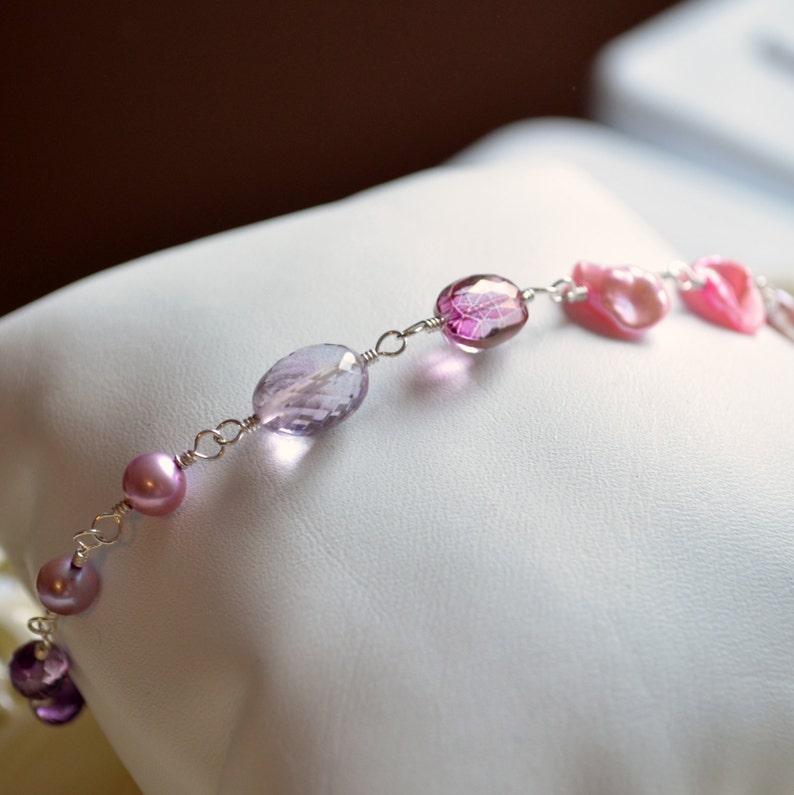 May include: A delicate bracelet featuring a mix of pink and purple oval-shaped gemstones and pearls, connected by silver wire.