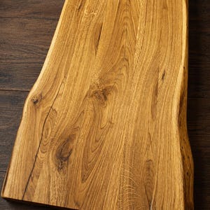 Può includere: Un tagliere rettangolare in legno caratterizzato da un bordo naturale su entrambi i lati. La superficie presenta un evidente motivo a venature color marrone dorato con nodi visibili e texture organiche su tutta la struttura in legno.