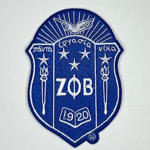 May include: A shield-shaped embroidered patch in royal blue and white. The design features the Greek letters ZΦB, a dove, three stars, two torches, and an open book with the year 1920. Greek text is inscribed above the letters in white thread.