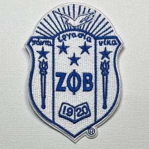 May include: A shield-shaped embroidered patch in white and blue featuring the Greek letters ZΦB, a dove, three stars, two torches, and an open book with the year 1920. The design includes Greek text at the top and a registered trademark symbol.