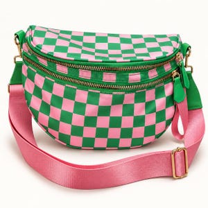 May include: A crescent-shaped crossbody bag featuring a vibrant pink and green checkered pattern. The bag includes two gold-toned zippered compartments and an adjustable pink nylon strap with gold-colored metal hardware and buckles.