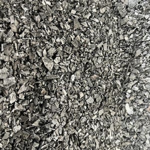 Könnte beinhalten: Eine dichte Ansammlung kleiner, unregelmäßiger, dunkelgrauer und schwarzer Holzkohlefragmente. Die Stücke variieren in Größe und Form, weisen scharfe, gezackte Kanten sowie eine matte, poröse Textur auf, die ein detailliertes, monochromatisches Muster bildet.