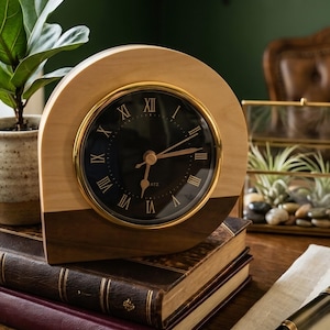 May include: A semi-circular desk clock featuring a two-tone light and dark wood frame, a black circular face with gold Roman numerals, and gold-colored hands. The clock is positioned on top of a stack of leather-bound books.
