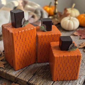 May include: Three rectangular wooden blocks painted orange with a textured surface, each topped with a dark brown square wooden block as a stem and a small green leaf accent. The blocks vary in height and are arranged in a cluster on a wooden surface.