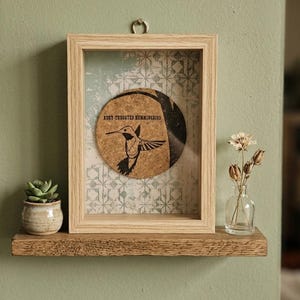 Può includere: Una cornice rettangolare a cassetta in legno chiaro contenente un inserto circolare in sughero con una grafica nera di un colibrì e il testo "RUBY-THROATED HUMMINGBIRD". La cornice presenta uno sfondo con motivi geometrici azzurri e bianchi.