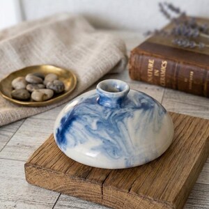 May include: A low-profile ceramic vase with a rounded, disc-like shape and a narrow neck. The surface features a marbled pattern of white and deep blue swirls, creating a fluid, cloud-like appearance across the entire exterior of the vessel.