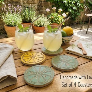 May include: A set of four round wooden coasters featuring a carved citrus slice design in yellow and green. Two coasters are green, one is yellow, and one is green, displayed alongside two glasses of lemonade with metal straws and a lemon.