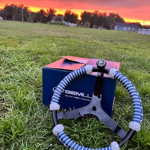 May include: A black boat steering wheel with a three-spoke design and a blue and white cord-wrapped rim. The wheel features a central knob and is positioned in front of a blue cardboard box with the text GEMLUX printed in white on the side.