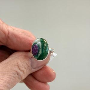 May include: Oval-shaped ring with a silver band. The ring features a large, polished gemstone with a mix of green, white, and deep red colors. The gemstone is set in a silver bezel.