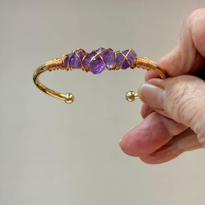 May include: Gold-toned cuff bracelet featuring raw amethyst stones. The purple stones are wire-wrapped with copper wire and set in a gold-colored metal band. The bracelet has rounded ball ends.