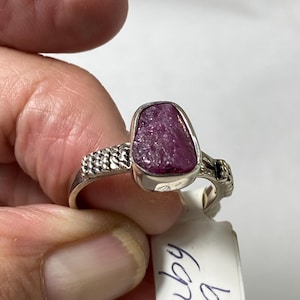 May include: A silver ring featuring a raw, deep pink ruby gemstone. The ring has a textured band and a bezel setting that holds the irregularly shaped stone. The tag next to the ring says "Ruby a".