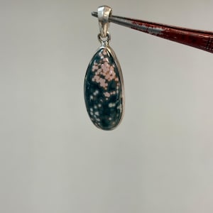 May include: A teardrop-shaped pendant with a silver-colored bail. The pendant features a dark green stone with small pink and white spots, set in a silver bezel. The pendant is held by a pair of tweezers.