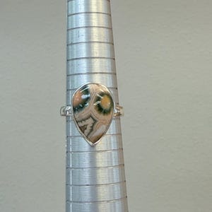 May include: A silver ring with a teardrop-shaped stone. The stone features a pattern of green, beige, and brown hues. The ring is displayed on a silver ring sizer.