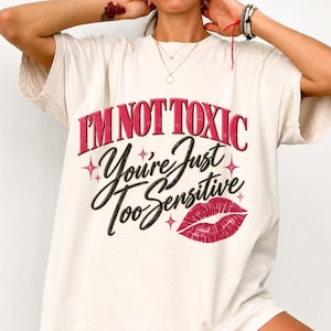 May include: An off-white short-sleeve t-shirt featuring the text I'M NOT TOXIC You're Just Too Sensitive in pink and black lettering. The design includes small pink star accents and a detailed graphic of a pair of red lips on the lower right.