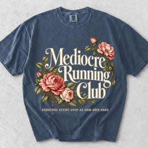 May include: A slate blue short-sleeve t-shirt featuring a graphic print of pink roses and green leaves surrounding the cream-colored text "Mediocre Running Club" and the smaller text "ENJOYING EVERY STEP AT OUR OWN PACE" centered below.