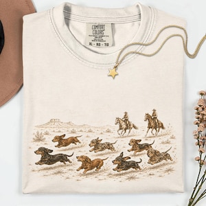 May include: A cream-colored t-shirt featuring a graphic print of several dachshunds running across a desert landscape with two cowboys on horseback in the background. The shirt has a Comfort Colors label at the neck and a gold star pendant necklace.