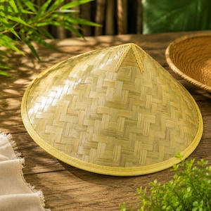 Puede incluir: Un sombrero cónico hecho de tiras de bambú trenzadas de color tostado claro, con una parte superior puntiaguda y un borde de tela amarilla reforzada a lo largo de la ala circular. El sombrero muestra un patrón de tejido diagonal.