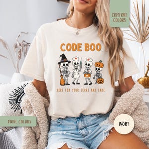 May include: An ivory short-sleeve t-shirt featuring a graphic of four skeletons in costumes with the text CODE BOO and HERE FOR YOUR SCARE AND CARE. The skeletons are dressed as a witch, a nurse, a medical professional, and a pumpkin character.