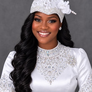 May include: A white cap adorned with intricate lace, clear crystals, and white fabric flowers, featuring a long, thin, pearl-accented stem extending from the side. The cap is paired with a matching white satin long-sleeve top with crystal embellishments.