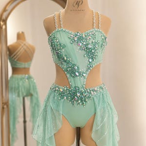 May include: A mint green dance costume featuring a sweetheart neckline, double shoulder straps, and cut-out sides. The bodice and waist are adorned with iridescent crystals, beads, and lace appliqués. A sheer, ruffled skirt overlay with sequin accents completes the ensemble, which is displayed on a mannequin labeled ap+ MOVEMENT.