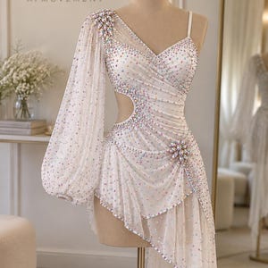 May include: A white asymmetrical dress featuring a single long sheer bishop sleeve, a cutout at the waist, and a draped skirt. The garment is embellished with iridescent crystals and a large floral-shaped crystal brooch at the hip.