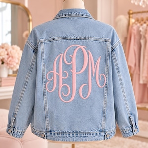 May include: A light blue denim jacket featuring a classic collar, long sleeves with buttoned cuffs, and a large, embroidered monogram on the back. The monogram consists of the letters APM in a decorative, light pink script font.