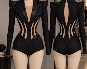 May include: A black bodysuit featuring a blazer-style collar, long sleeves, and structured shoulders. The garment includes sheer mesh panels at the waist and torso, embellished with small black studs. The back displays a high collar and cutout.