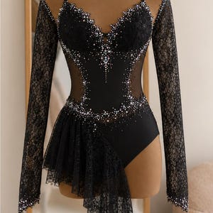 May include: A black leotard featuring long lace sleeves, a sweetheart neckline, and sheer mesh side panels. The bodice is embellished with iridescent crystals along the neckline and waist. An asymmetrical lace skirt overlay hangs from the right hip.