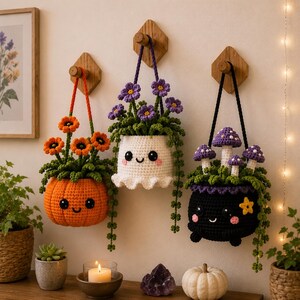 May include: Three crocheted hanging planters featuring cute faces. The left is an orange pumpkin with orange flowers, the centre is a white pot with purple flowers, and the right is a black cauldron with purple mushrooms and a yellow star.