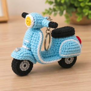 Puede incluir: Un llavero de scooter tejido a ganchillo en color azul claro con asiento negro, neumáticos negros con centros grises, un faro amarillo y una pequeña luz trasera roja. El scooter está hecho de hilo e incluye una anilla de metal plateado.