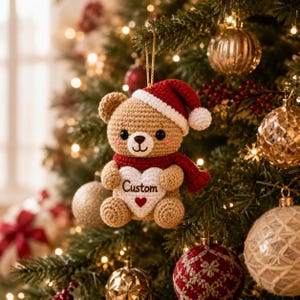 Può includere: Un ornamento a forma di orsetto all'uncinetto color marrone chiaro, con un cappello da Babbo Natale rosso e bianco e una sciarpa rossa. L'orsetto tiene un cuore bianco con la scritta Custom ricamata in nero sopra un piccolo cuore rosso.