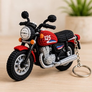 Puede incluir: Un llavero de motocicleta tejido a ganchillo con un depósito de combustible rojo con el número 125 en blanco, un asiento negro, manillar negro y un tubo de escape plateado. La moto tiene un faro redondo blanco, intermitentes naranjas y una argolla.