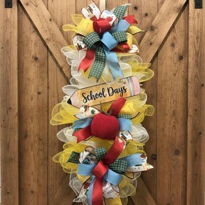 May include: A teardrop-shaped wreath made of yellow and white mesh, featuring red, blue, and green plaid ribbons. A wooden sign shaped like a pencil with the text School Days is centered, along with a red apple accent in the middle of the arrangement.