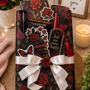 May include: A gift set wrapped in black paper with a red rose pattern, featuring a white satin bow, a black pen, a red pen, a Bigelow tea bag, a butterfly sticker, a stack of books sticker, and a bookmark with the text Just one more chapter.