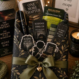 May include: A gift set wrapped in black paper with gold leaf patterns, tied with an olive green ribbon and a gold heart charm. It contains a pen, a Bigelow green tea bag, and several bookmarks and stickers featuring book-themed text and illustrations.