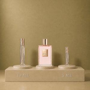 Op de afbeelding: Een rechthoekige parfumfles met lichtroze vloeistof en een gouden dop, geflankeerd door twee doorzichtige glazen verstuivers met gouden doppen. De items staan op een beige platform met de tekst 10 ML en 5 ML gegraveerd op de voorzijde.