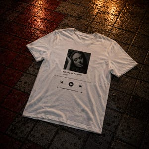 Op de afbeelding: Een wit t-shirt met korte mouwen en een zwart-witte grafische print van een muziekspeler-interface. Het ontwerp bevat een portret van Adele, de tekst Set Fire to the Rain, de artiestennaam Adele en standaard afspeelbedieningspictogrammen.