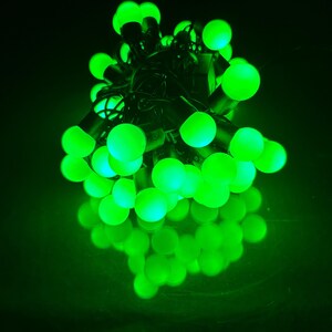 May include: A cluster of glowing green spherical LED string lights arranged in a dense, tangled bundle. Each small, round bulb emits a bright, vibrant green light, creating a concentrated orb of illumination against a dark, black background.