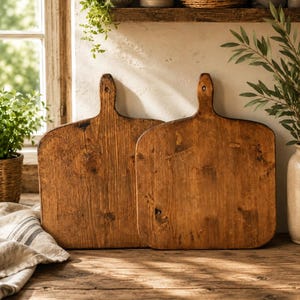 May include: Two rectangular wooden cutting boards with rounded corners and a vertical handle at the top center. The boards feature a medium brown wood grain with visible knots and a smooth, flat surface, standing upright side by side.