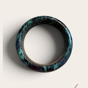 May include: A circular bangle bracelet featuring a polished surface with a marbled pattern of deep forest green, black, and hints of dark magenta. The smooth, rounded band has a glossy finish that reflects light, showcasing the natural colour variations.