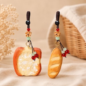May include: Two hanging ornaments featuring carved wooden bread shapes. One is a square slice of toast with a textured surface, and the other is a long baguette. Both are suspended by dark brown braided cords with red, green, and flower beads.