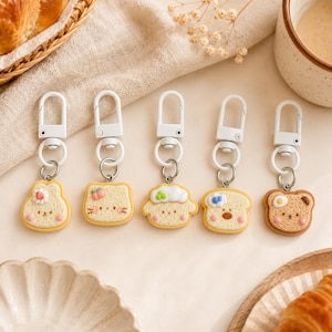 May include: Five keychains featuring miniature bread-shaped charms with various toppings including a strawberry, peach, green garnish, blueberry, and fried egg. Each charm has a smiling face and is attached to a white swivel lobster clasp.