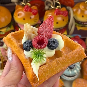 Puede incluir: Un dije con forma de gofre triangular que presenta una superficie texturizada de color marrón dorado espolvoreada con polvo blanco. Está cubierto con una porción de crema blanca, una rodaja de fresa roja, una frambuesa roja, dos arándanos oscuros y una pequeña ramita verde.