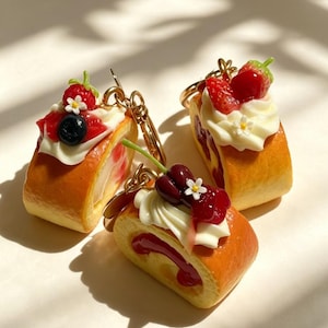 Pode incluir: Três porta-chaves em miniatura em forma de torta suíça com pão de ló dourado, redemoinhos de creme branco e coberturas de frutas, incluindo morangos, mirtilos e cerejas, decorados com pequenas flores brancas e argolas douradas.