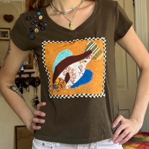 May include: A brown V-neck t-shirt featuring a square patch on the chest with a multi-colored fabric fish appliqué. The fish is composed of various patterned fabrics in shades of blue, brown, and white. A cluster of assorted buttons adorns the right shoulder.