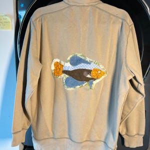 May include: A beige long-sleeve sweatshirt featuring a large, centered fish appliqué. The fish is constructed from various fabric patches in shades of blue, brown, and yellow, with a small white button serving as the eye of the fish.