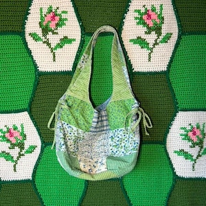 May include: A hobo-style shoulder bag crafted from a patchwork of various green, blue, and white fabrics featuring floral, gingham, and plaid patterns. The bag includes two side drawstrings with fabric ties and a single integrated shoulder strap.